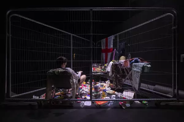 The colonialism station, depicting a person sat inside a fenced-off triangle. There is an England flag and lots of rubbish and objects scattered across the floor.