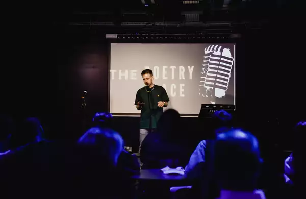 A poet performs spoken word poetry on stage in front of an audience, with a projection reading "The Poetry Place" behind him.