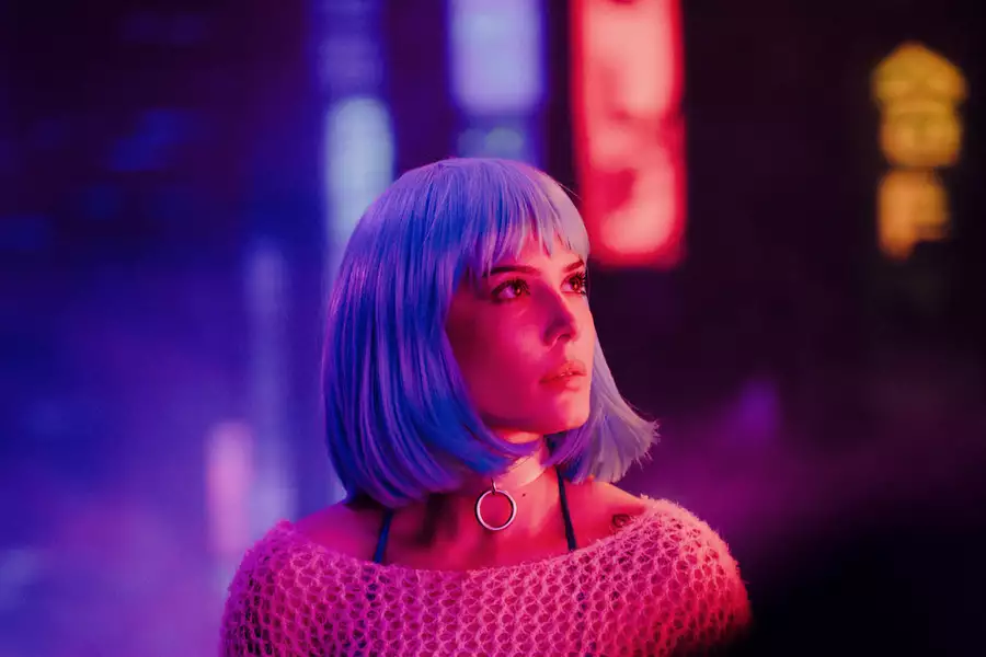 Halsey with a short, straight, light blue wig and wearing a silver ring choker looks upward under neon purple and pink lights in a futuristic, cyberpunk-style setting.