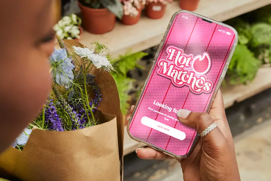 A person holding a bouquet of flowers is using a smartphone displaying the "Hot Matches" dating app. The app screen features a bright pink background with the tagline “Looking for love?” and a button labeled “Join Now.”