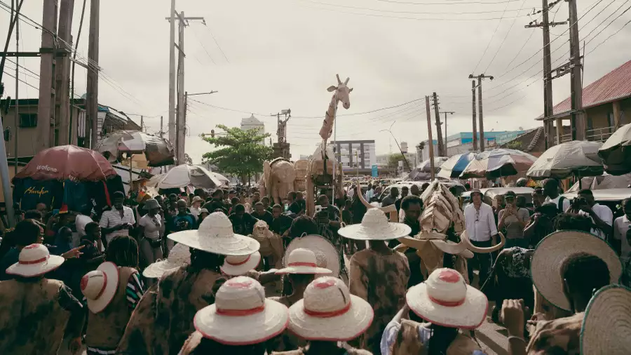 A large crowd of people at an outdoor market wearing hats. They are looking at large giraffe and elephant puppets.