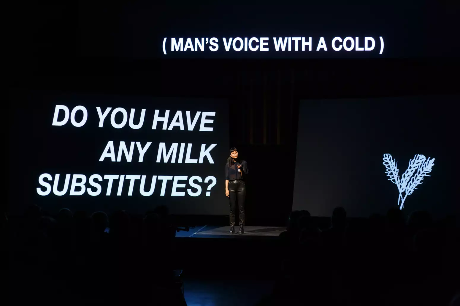 The artist Christine Sun Kim stands on a stage in the centre of a dark room. A screen above her shows in white text "(MAN'S VOICE WITH A COLD)", another to the left reads "DO YOU HAVE ANY MILK SUBSTITUTES?" and to the right is a white line drawing of oats.