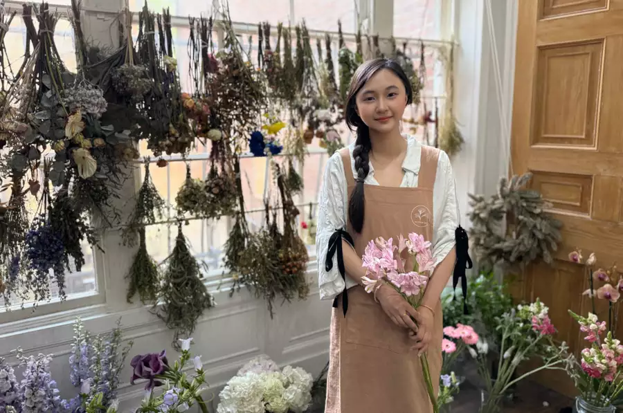 Kiki smiling in her art studio, wearing a light brown apron, holding flowers. There are many bunches of flowers behind her.