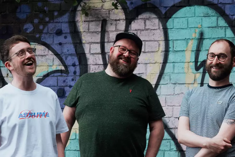 Three members of Kiss Me Again smiling and laughing. They are stood in front of a graffiti covered wall.