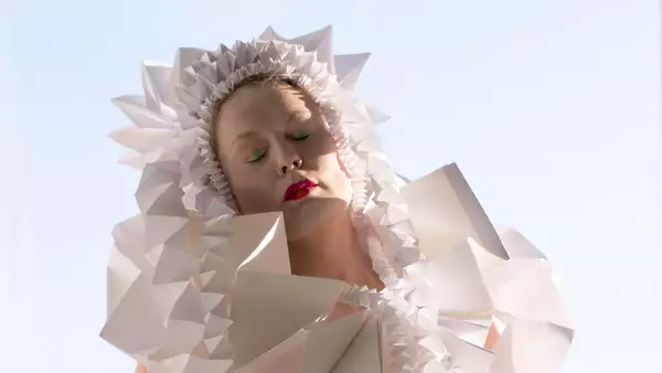 A person poses in an avant-garde wearable art piece made of intricate folded white paper structures. Their eyes are closed, with bold makeup, and they stand in front of a blue sky.