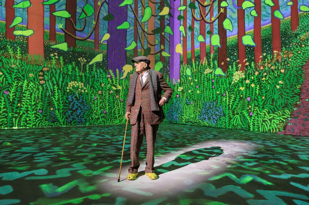 A man in a brown suit and flat cap holding a walking stick standing in a room with bright paintings projected on the walls and floor