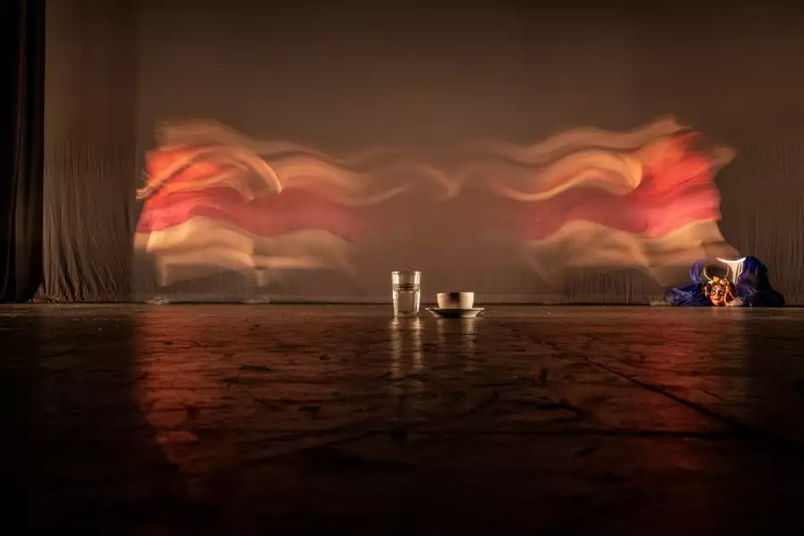 A glass of water and cup and saucer are placed on the floor. Behind them is a curtain and to the right is a puppet.