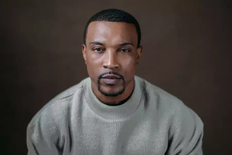 Headshot of Ashley Walters, wearing an off-white jumper. The background of the photo is brown.
