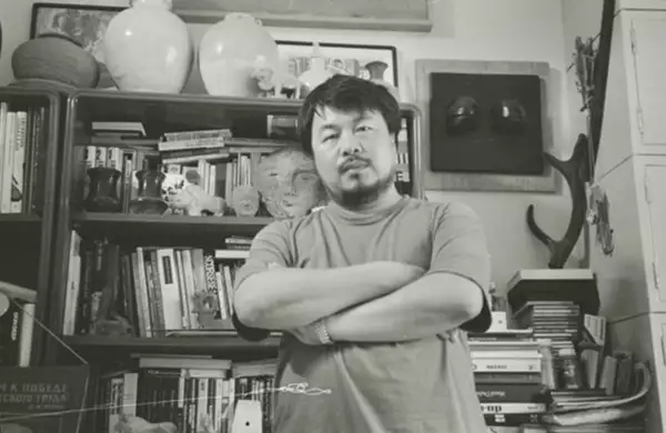 Black and white photograph of Ai Weiwei stood in front of a bookshelf
