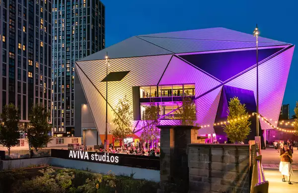 A photo of the venue Aviva Studios lit up with purple lights with crowds enjoying Festival Square by the river