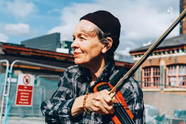 A photograph of Laurie Anderson stood outside at a construction site, plucking a string of an electric violin which is resting on her left shoulder,