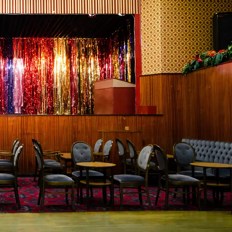 A pub stage with a glittery curtain, blue chairs, tables and a patterned carpeted floor.