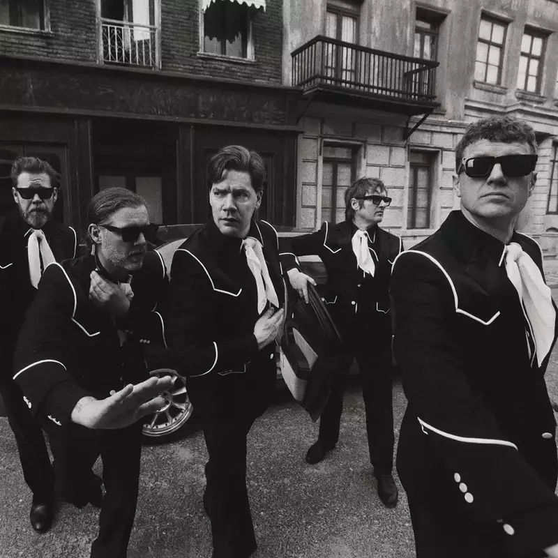 Black and white photograph of the five members of The Hives wearing matching suits and sunglasses striking dynamic poses on a city street. There is a car in the background.