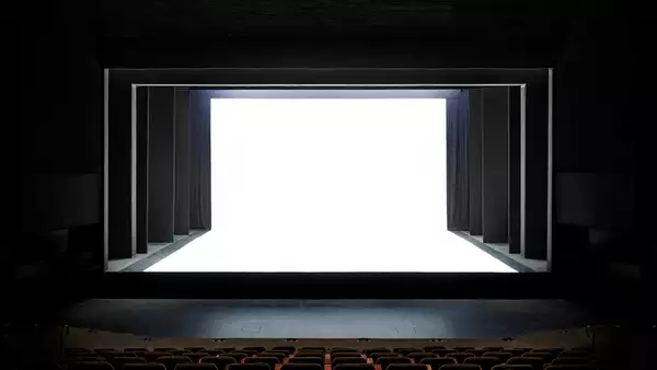 A theater stage with a dramatic, modern design is shown, featuring black framing and a stark white, illuminated backdrop. The contrast between the dark seating area and the bright stage creates a striking visual effect.