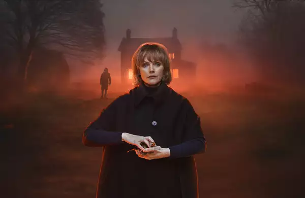 The Actor Maxine Peake clutching a robin and looking directly at the viewer. She is stood in front of a house with an orange eerie glow.