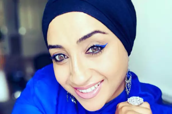 A close-up of a woman wearing a blue shirt and headscarf and silver earrings. She is looking up at the camera smiling.