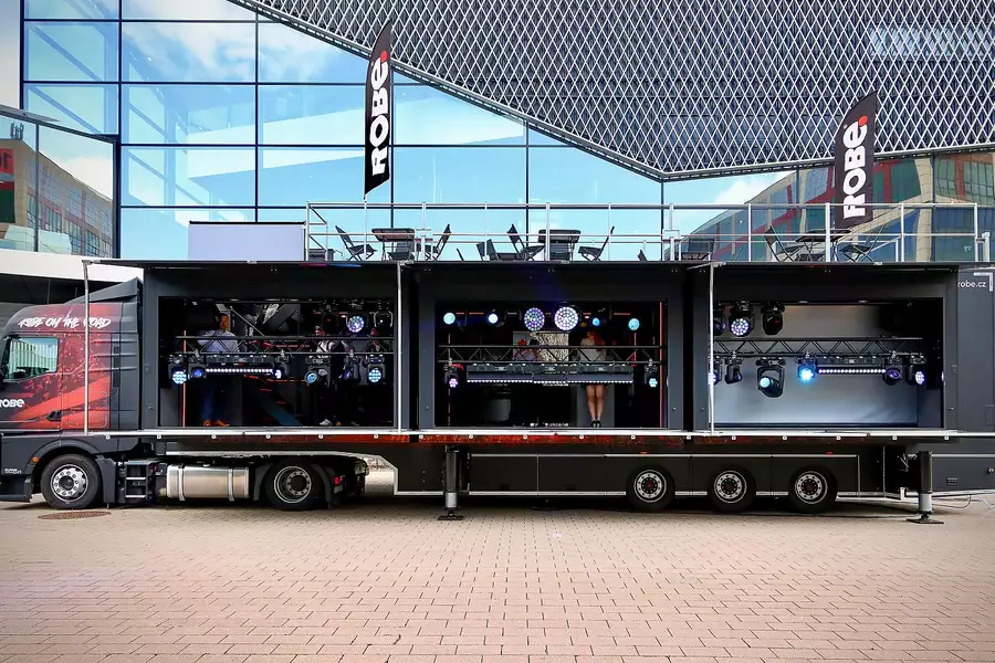A large black semi-truck trailer is transformed into a mobile stage display featuring professional lighting equipment and a small viewing area inside. The setup is branded with “ROBE” flags and includes a rooftop seating area above the trailer.
