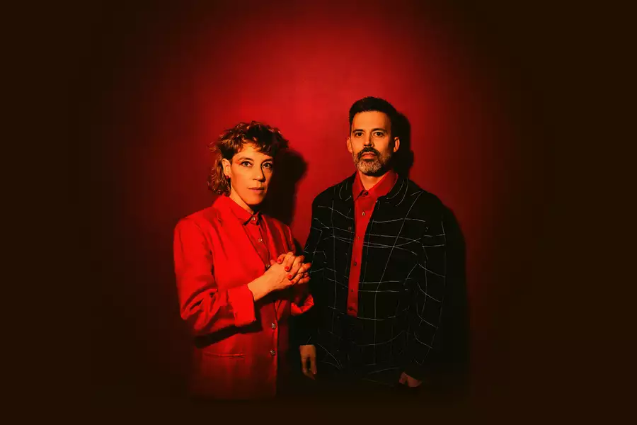 Two members of tune-yards wearing suits on a red background. The image features vibrant colours and dramatic red lighting.