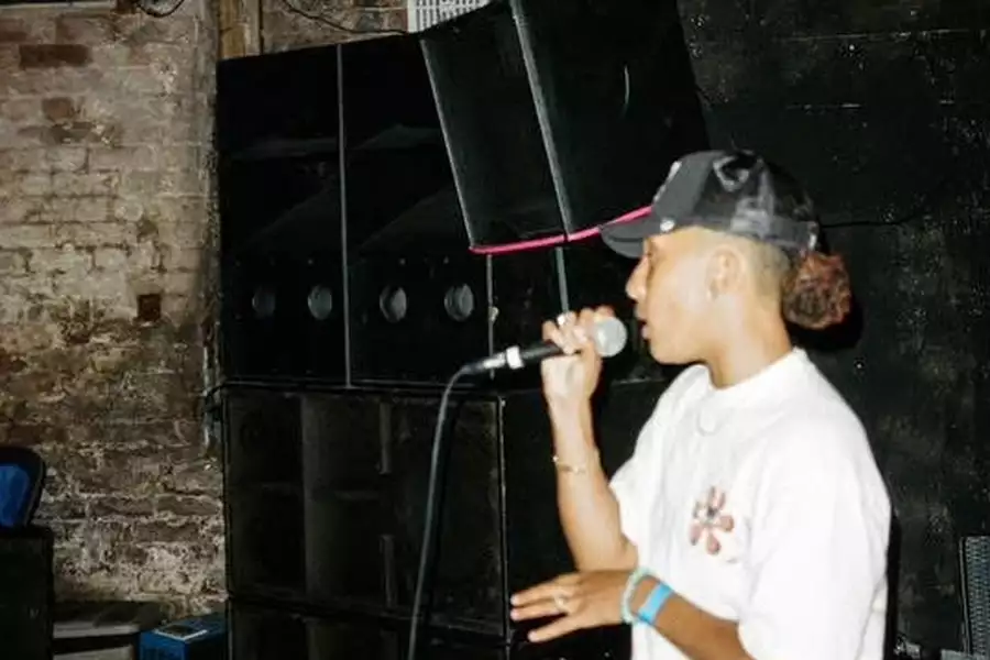 True Aubz performing into a microphone wearing a black cap. They are stood in front of a big sound system.