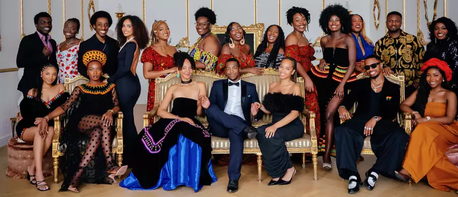 A large group of people pose together in a luxurious room with gold-trimmed walls and ornate furniture. They wear a mix of modern formalwear and vibrant traditional African attire, smiling warmly at the camera.