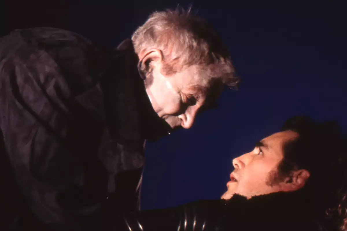 Russell Enoch towering over Mark Rylance, who is looking up at him in fear. Russell has his hands on Mark's neck. Both are wearing black coats.