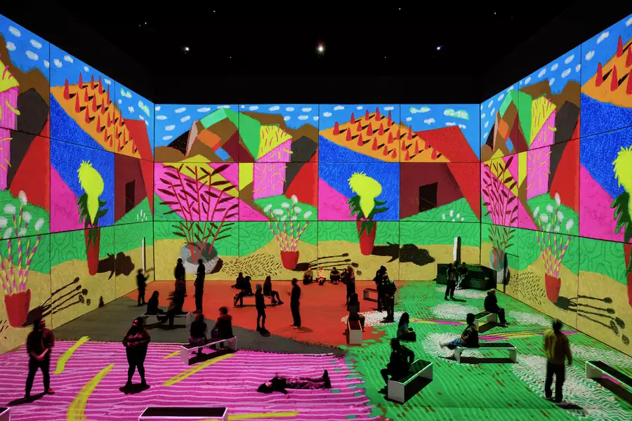 An image from David Hockney: Bigger & Closer (not smaller & further away), showing people in the exhibition seated and standing, surrounded by his artwork