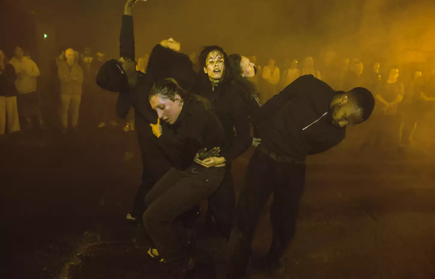 A photo of warehouse industrial space lit in yellow light with a group of dancers holding each other