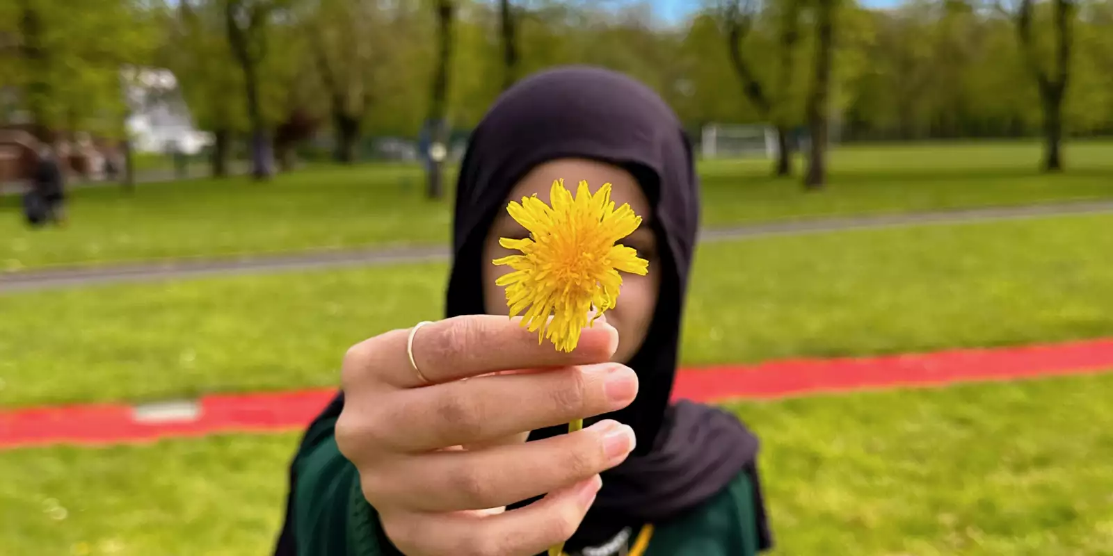 A girl stood in a park holding a dandelion in front of her face