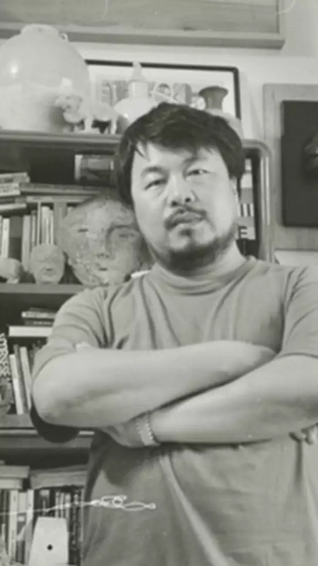 Black and white photograph of Ai Weiwei stood in front of a bookshelf