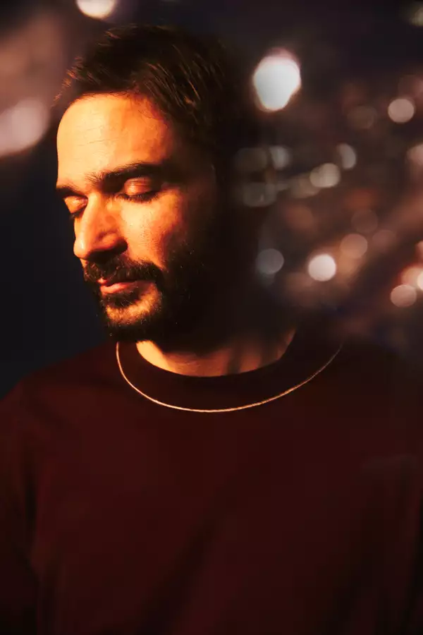 Headshot of Jon Hopkins with his eyes closed. He is wearing a maroon jumper and gold chain. The background shows blurry lights.