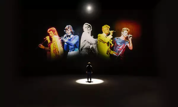 A person looking at five versions of David Bowie singing on a large screen