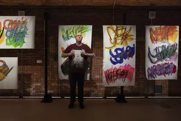 A photo of artist Michael Beard standing in front of a gallery wall of his paintings which feature brightly coloured layers of paint used to write the names of cities in a repetitive pattern