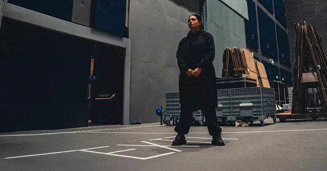 Hannah Laing dressed in black, standing in the Warehouse surrounded by equipment and stacked materials.