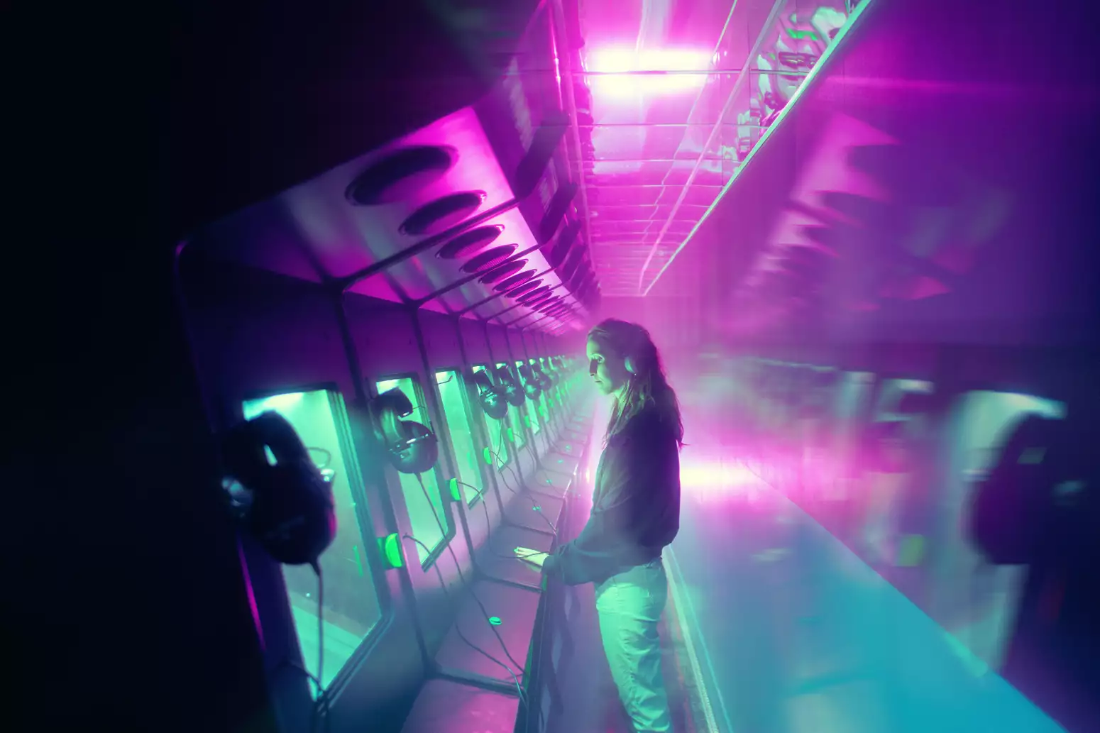 A person touching a video game station in ARCADE by Darkfield, wearing headphones. There are several video game stations. The lighting is blue and pink.