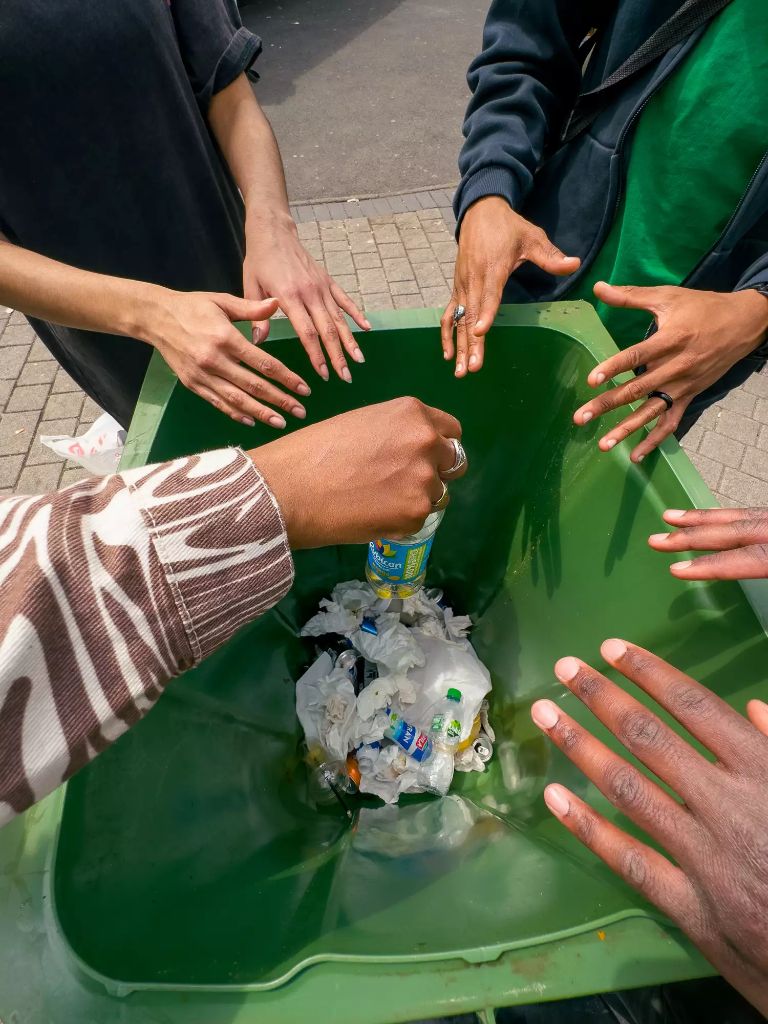 Several hands hover over a green bin filled with rubbish