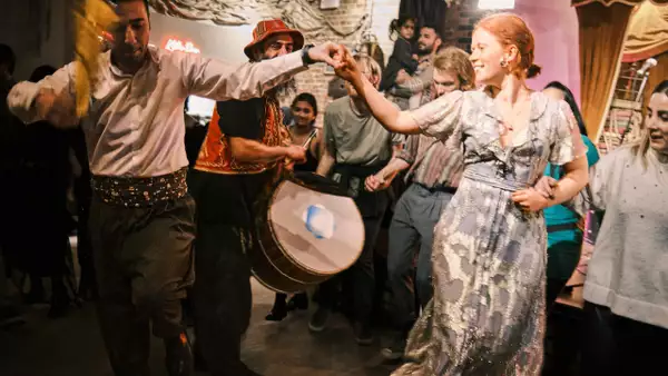 A group of people enjoy folk dancing indoors; two people hold hands while others watch and play traditional drums. The scene is lively and festive, with warm lighting and colourful costumes.