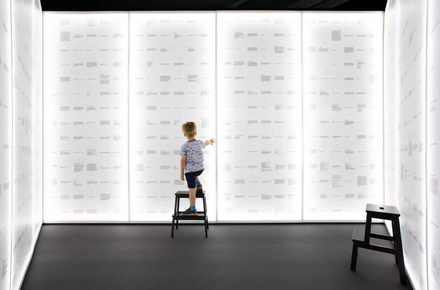 A photo of a little boy on a step looking at a brightly lit wall of messages from local school children about the future