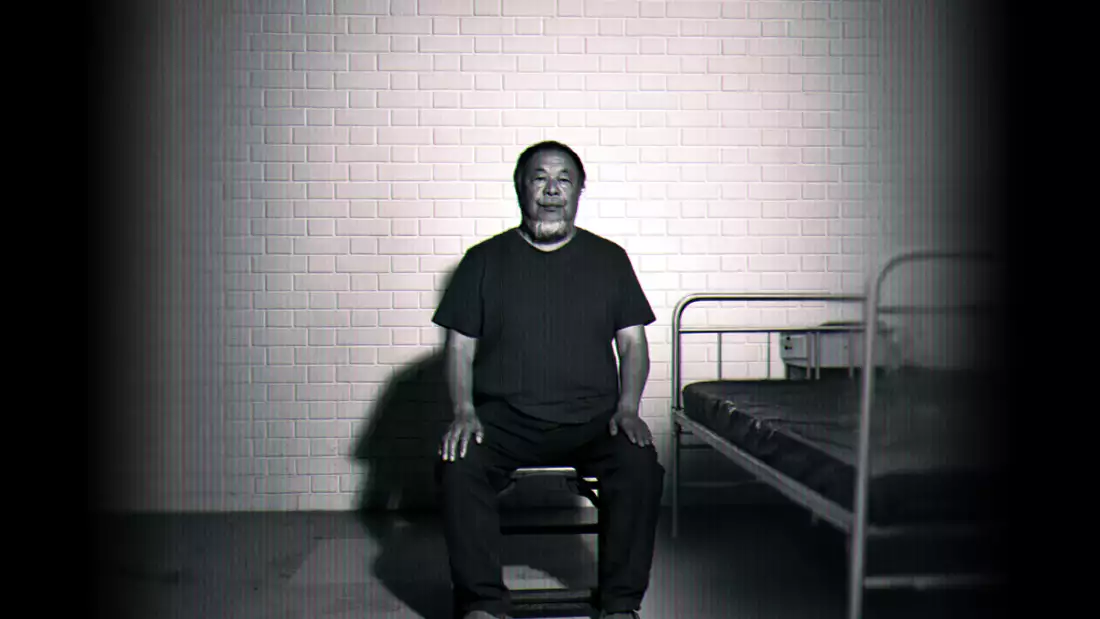 Black-and-white image of Ai Weiwei sitting on a chair beside a simple metal bed against a brick wall.