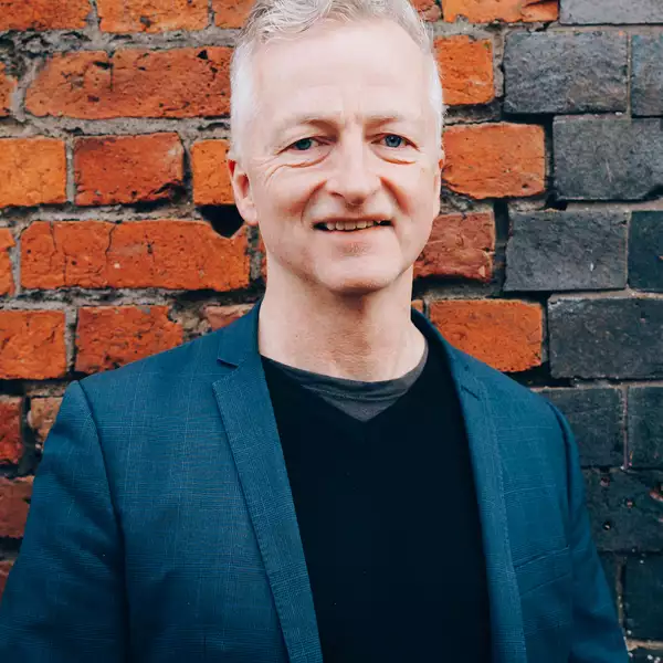 Headshot of John McGrath wearing a navy suit jacket and stood in front of a brick wall