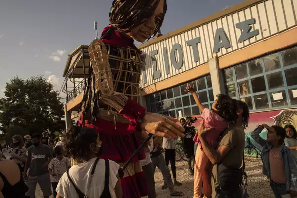 The Little Amal puppet looks down and reaches out to a toddler, held up by her parent. Behind her, another young girl shields her eyes from the sun with her hand and gazes up at Little Amal. In the background is a crowd including a cameraman.