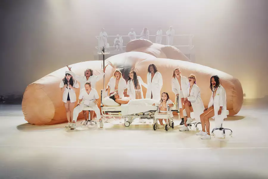 A group of performers on stage wearing white medical dress in front of a giant sculpture of a woman's body