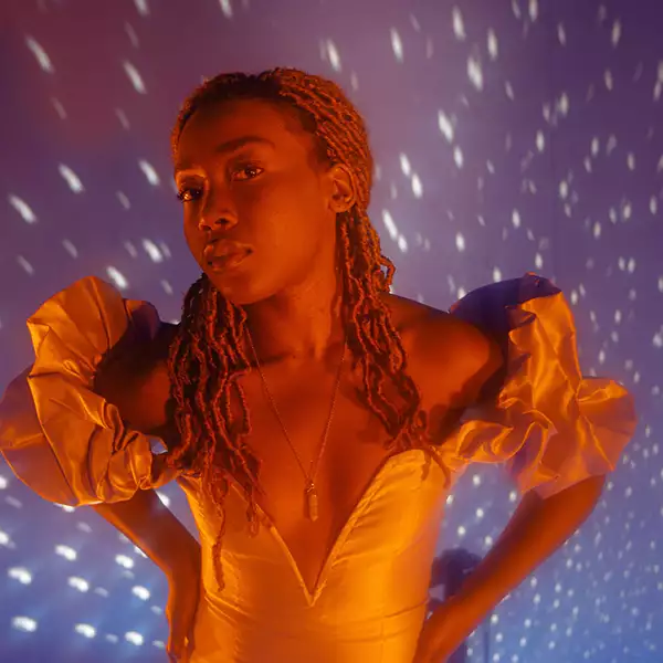 The artist Bitez with her head slightly tilted, eyes looking towards the viewer and her hands placed on her hips. Her hair is in light brown braids and she is wearing a silver dress with a deep V line which is puffed at the shoulders. She is wearing a plunging crystal necklace and the background shows shimmers from a disco ball.