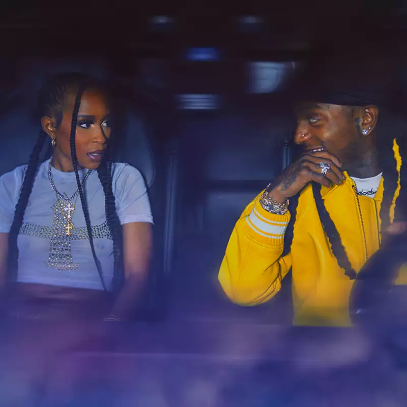 Jacquees and Dej Loaf sit in the front seats of a car, facing each other. Dej Loaf wears a light blue crop top with a bejewelled cross, while Jacquees wears a yellow hoodie and smiles warmly.
