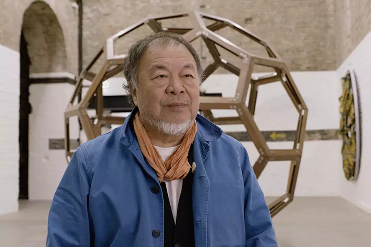 Ai Weiwei talking to the camera in his studio. Behind him is a large circular sculpture.
