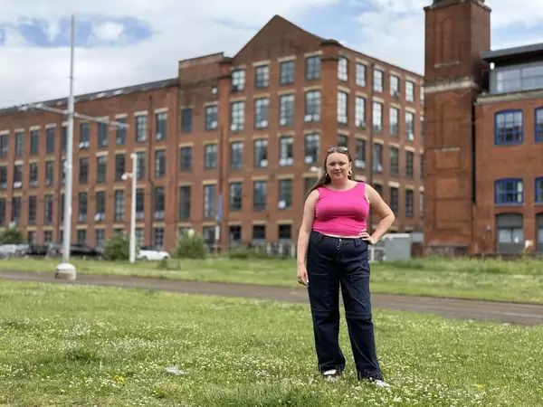 Ellie Brown wearing a bright pink top and jeans, stood outside a converted mill on the grass