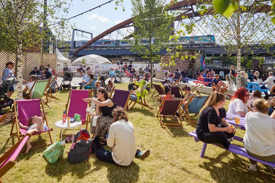 Factory Square filled with people sat on the grass and deckchairs, drinking and chatting. The sun is shining.