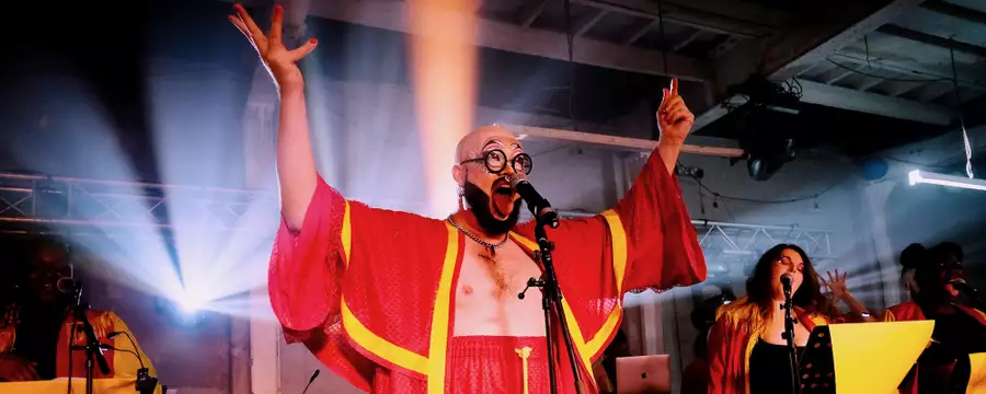 A performer in a bright red choir robe sings into a microphone with raised arms under dramatic stage lights, with backup singers behind them.