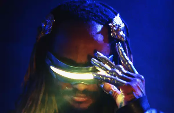 Thundercat wearing futuristic glowing visor glasses and metallic claw-like gloves against a dark blue background.