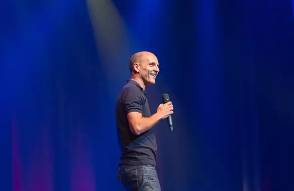 Carl Barron smiling and holding a microphone on stage, with blue curtains behind him