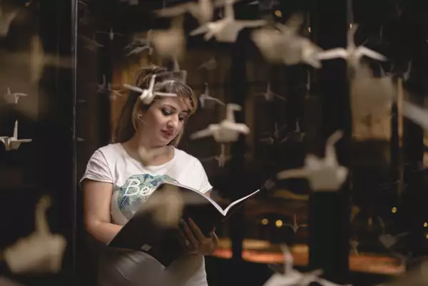 A woman in a white t-shirt reads from an A4 size book. She wears her hair in a high ponytail with a fringe. In the foreground there are paper cranes.